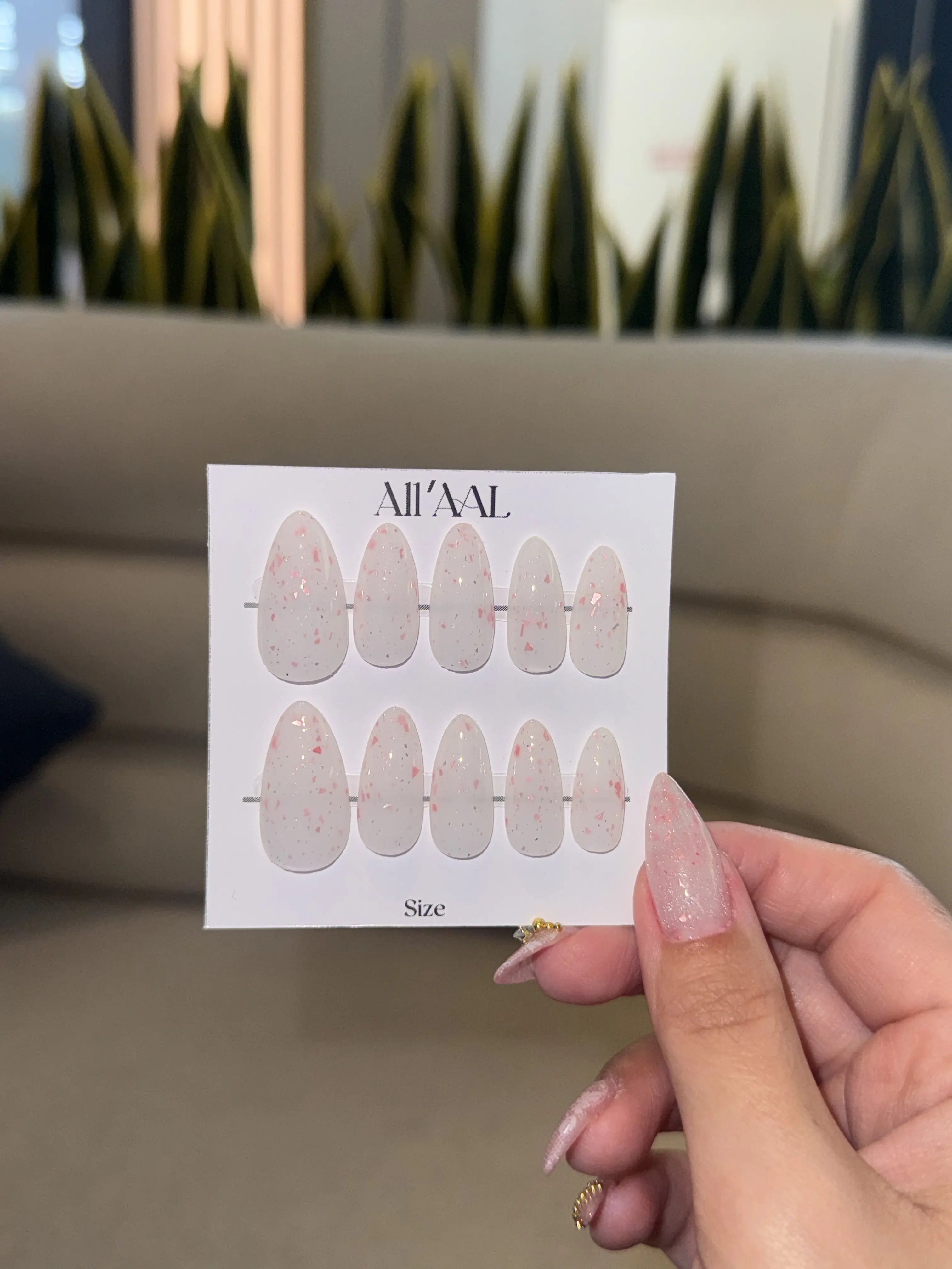 Hand holding a nail art design sheet with 'Alilaal' branding against a blurred indoor background.