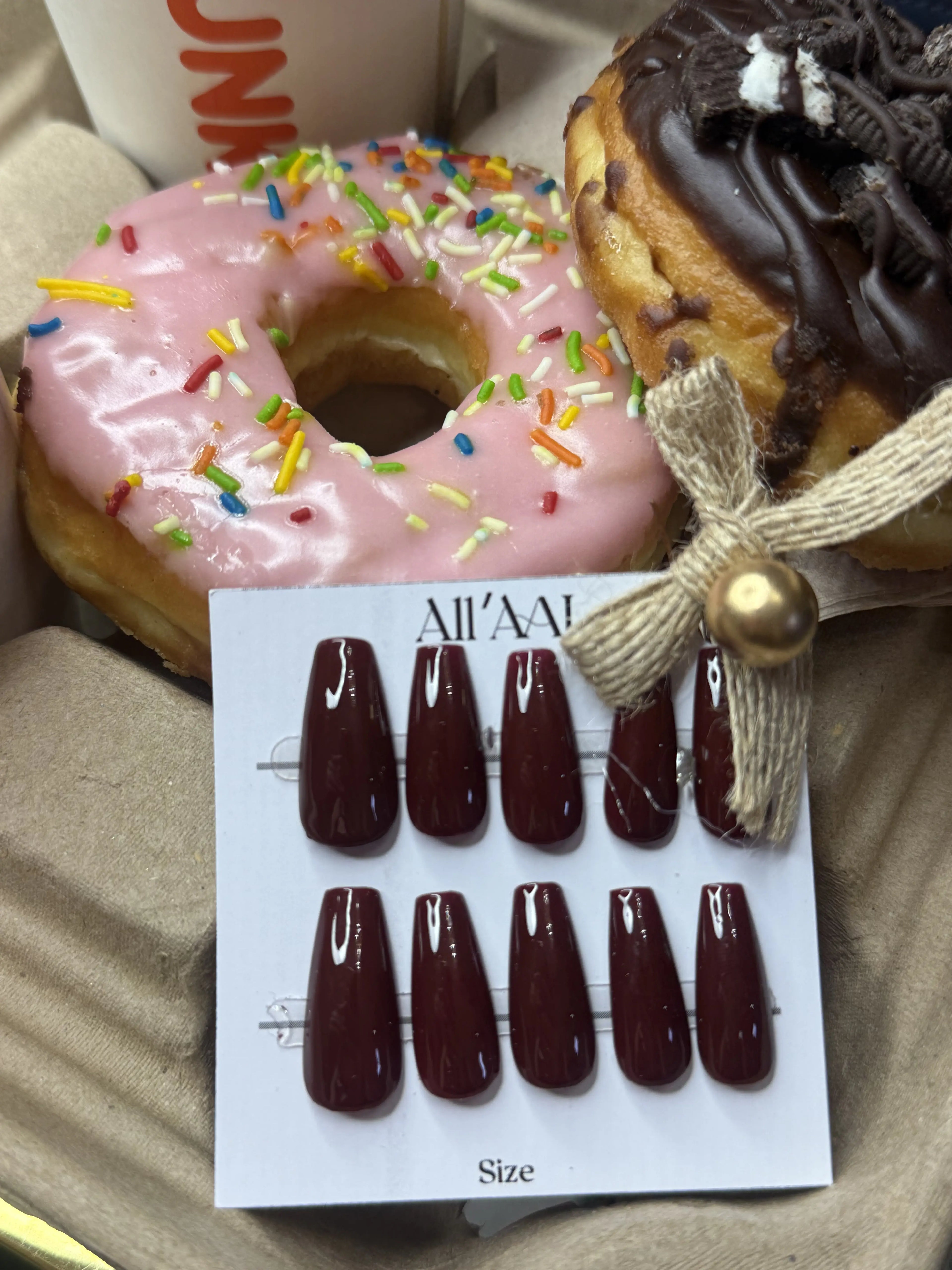 A set of glossy dark burgundy press-on nails displayed on a white card labeled 'All'AA' Size', styled with donuts and a coffee cup in the background.