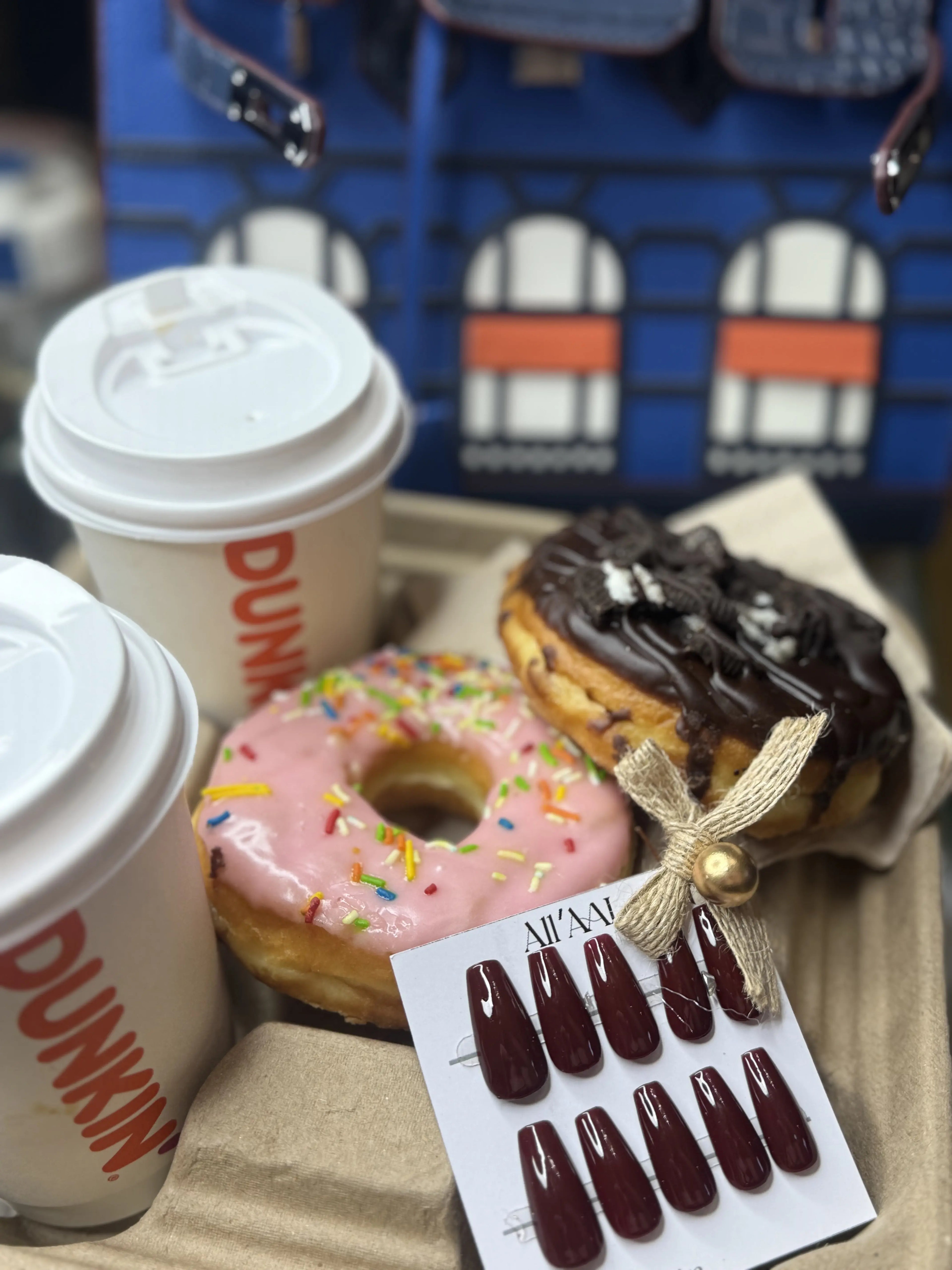 A tray of assorted pastries and coffee cups, featuring a pink sprinkle donut, a chocolate-topped pastry with a ribbon, two Dunkin' coffee cups, and a card displaying dark burgundy press-on nails.