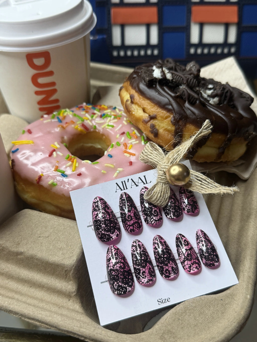 A set of decorative press-on nails on a white card labeled 'ALL'AL' is displayed in a tray with two donuts and a Dunkin' coffee cup in the background.