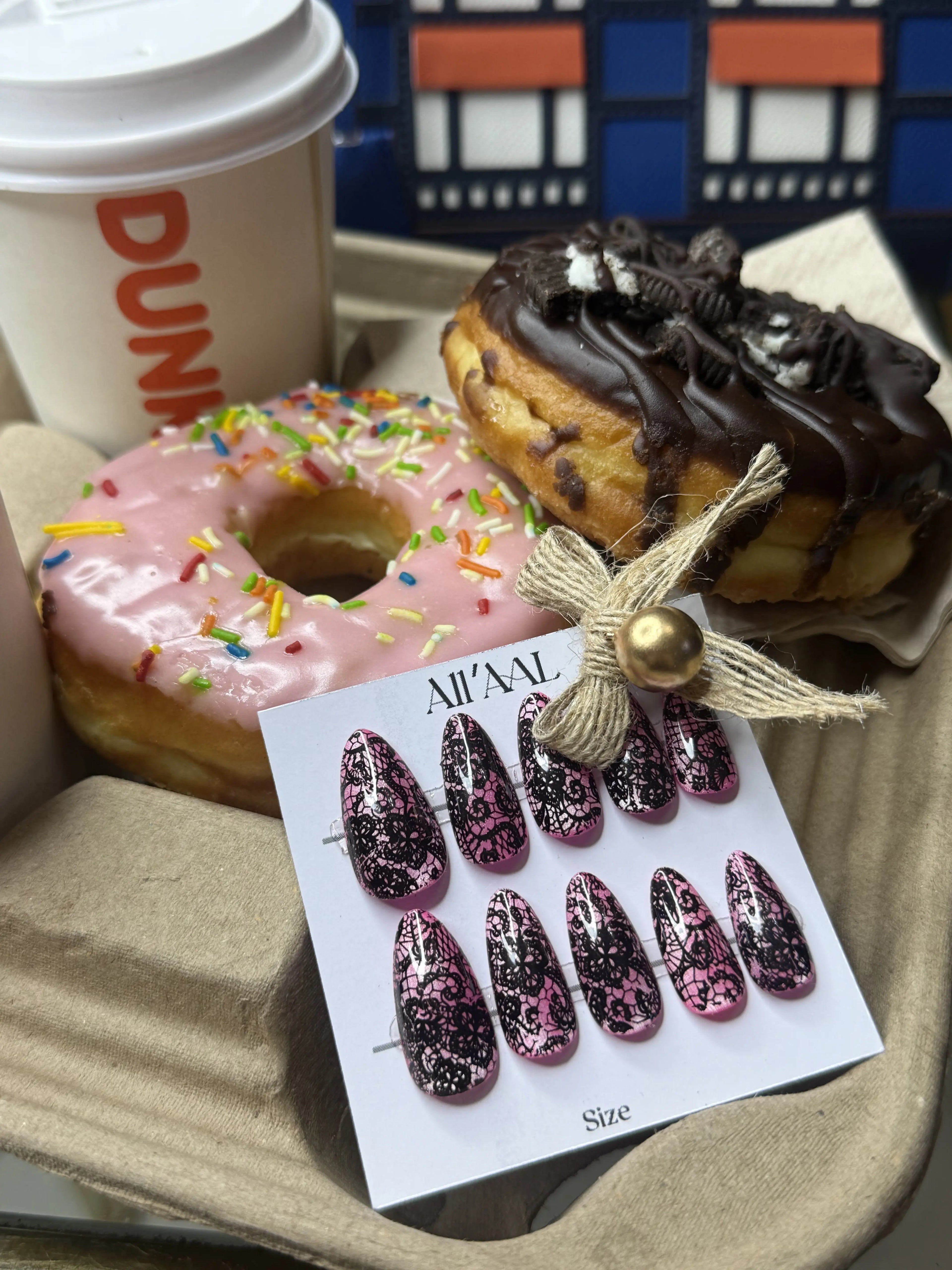 A set of decorative press-on nails on a white card labeled 'ALL'AL' is displayed in a tray with two donuts and a Dunkin' coffee cup in the background.