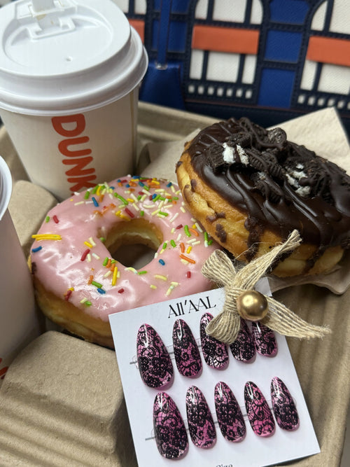 A tray of treats featuring a pink frosted donut with rainbow sprinkles, a chocolate-covered pastry topped with cookie crumbs, a Dunkin' coffee cup, and a card of press-on nails with a decorative twine bow.