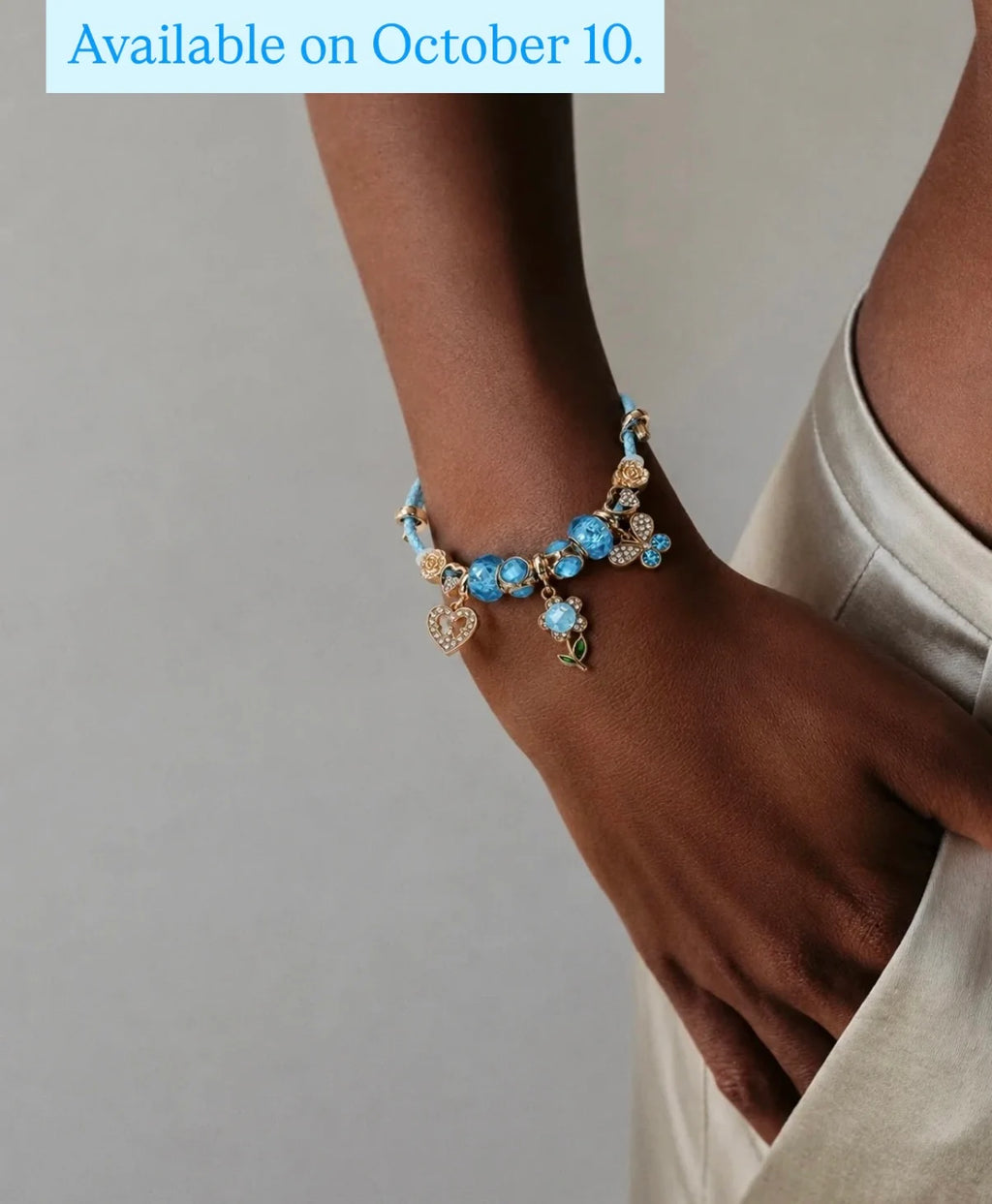 A close-up of a person's wrist and hand wearing a delicate charm bracelet with light blue beads, gold-toned floral details, and heart charms. A light blue text banner at the top reads 'Available on October 10.