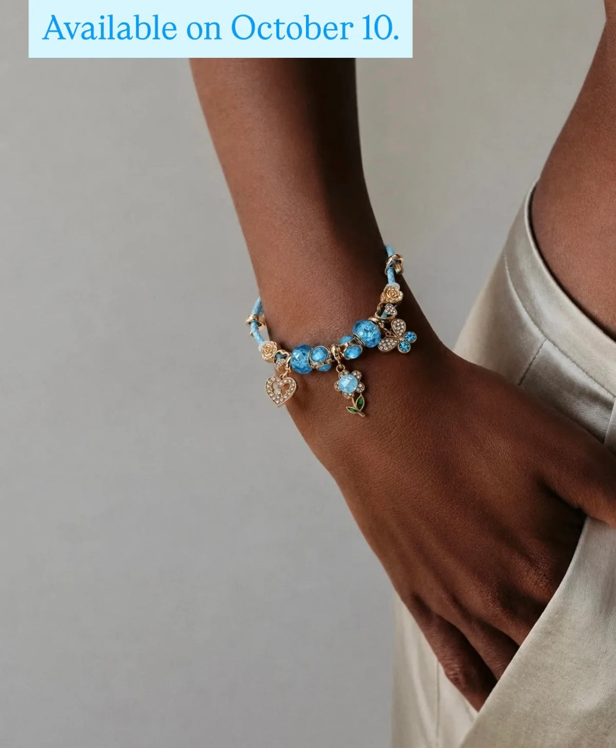 A close-up of a person's wrist and hand wearing a delicate charm bracelet with light blue beads, gold-toned floral details, and heart charms. A light blue text banner at the top reads 'Available on October 10.