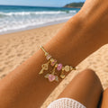 A gold charm bracelet with pink gemstone beads, a key charm, and a winged heart charm, shown worn on a person's wrist at a sunny beach with the ocean in the background.