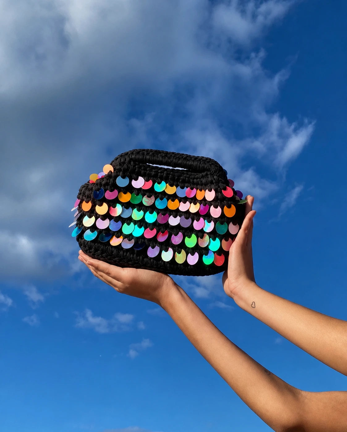 A black crocheted handbag covered in colorful sequins, held up by two hands against a bright blue sky with clouds.