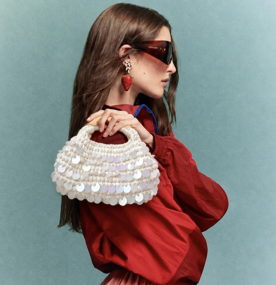 A woman in a red jacket and dark sunglasses holds a white beaded handbag decorated with silver disc embellishments, set against a solid teal background.