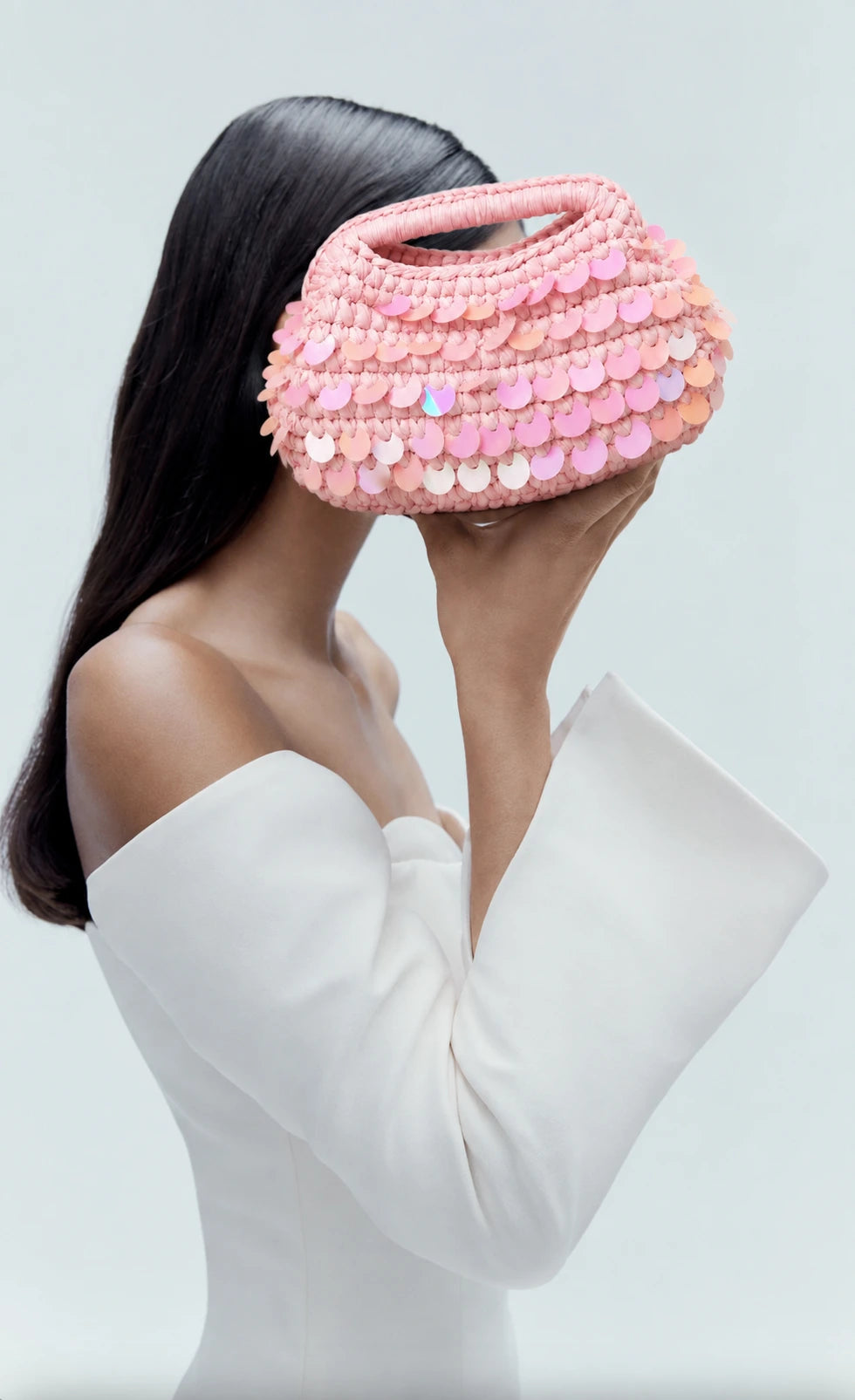 A woman in a white off-the-shoulder top holds a pink crocheted handbag in front of her face. The bag is covered in rows of pink and iridescent sequins and has a top handle, set against a clean, light gray background.