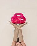 A bright pink crocheted handbag with iridescent sequins, held up by two crossed beige leather legs against a plain light beige background.
