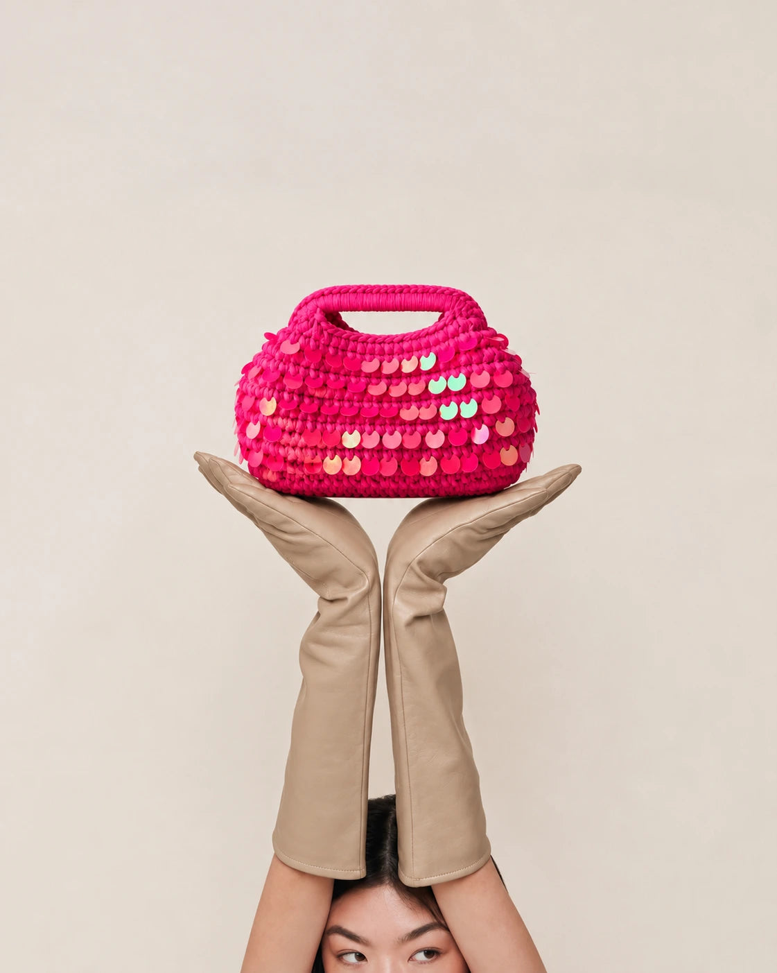 A bright pink crocheted handbag with iridescent sequins, held up by two crossed beige leather legs against a plain light beige background.