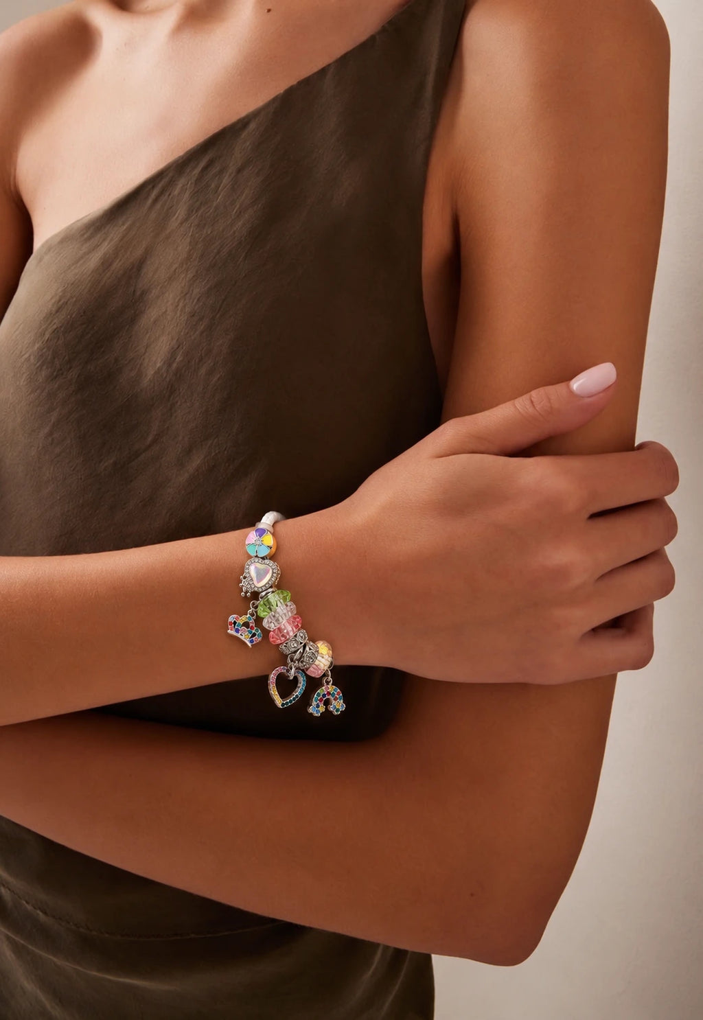 A close-up of a model wearing a silver charm bracelet with colorful heart charms, paired with a dark brown one-shoulder dress against a neutral beige background.