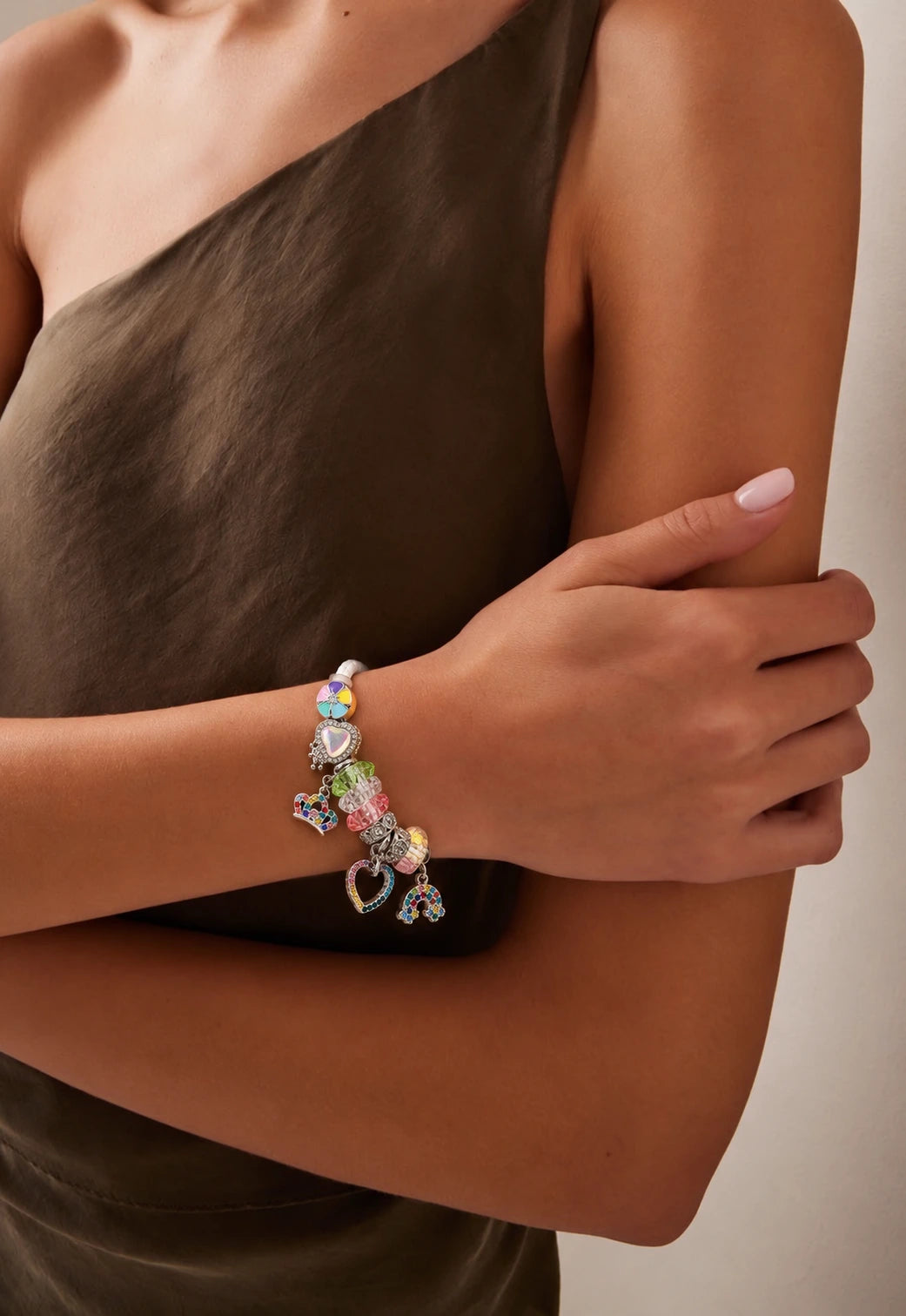 A close-up of a model wearing a silver charm bracelet with colorful heart charms, paired with a dark brown one-shoulder dress against a neutral beige background.