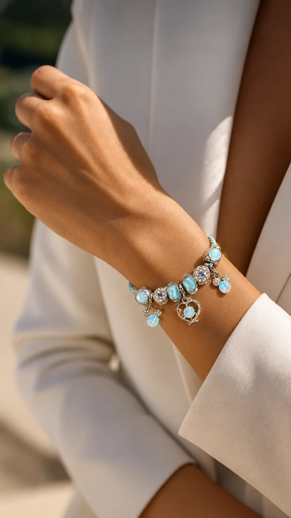 A close-up of a person's wrist wearing a delicate silver-toned charm bracelet with light blue oval charms and clear crystal accents, paired with a white blazer.