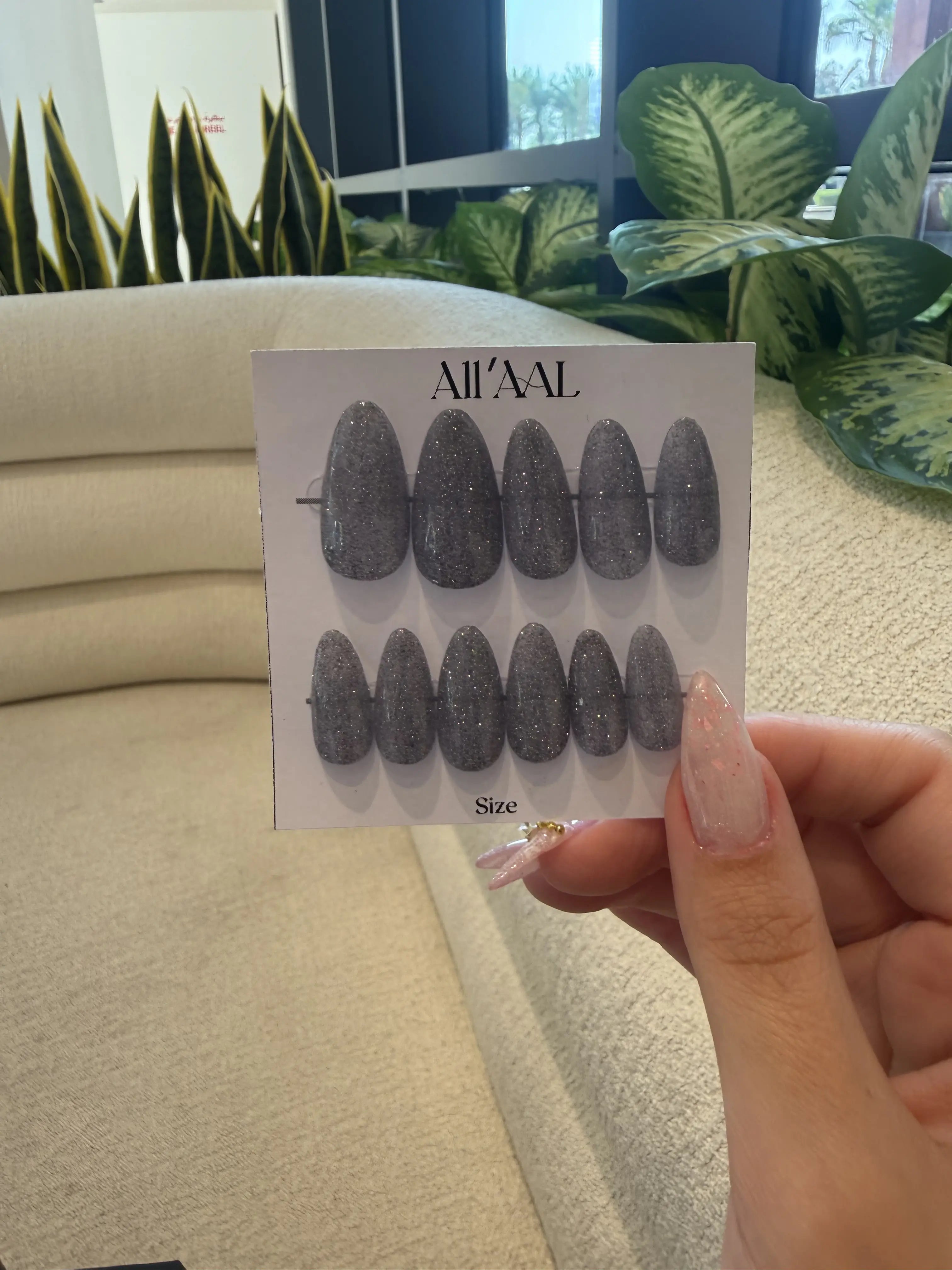 Card with glittery black nail art designs held by a hand in front of a beige sofa and plants.