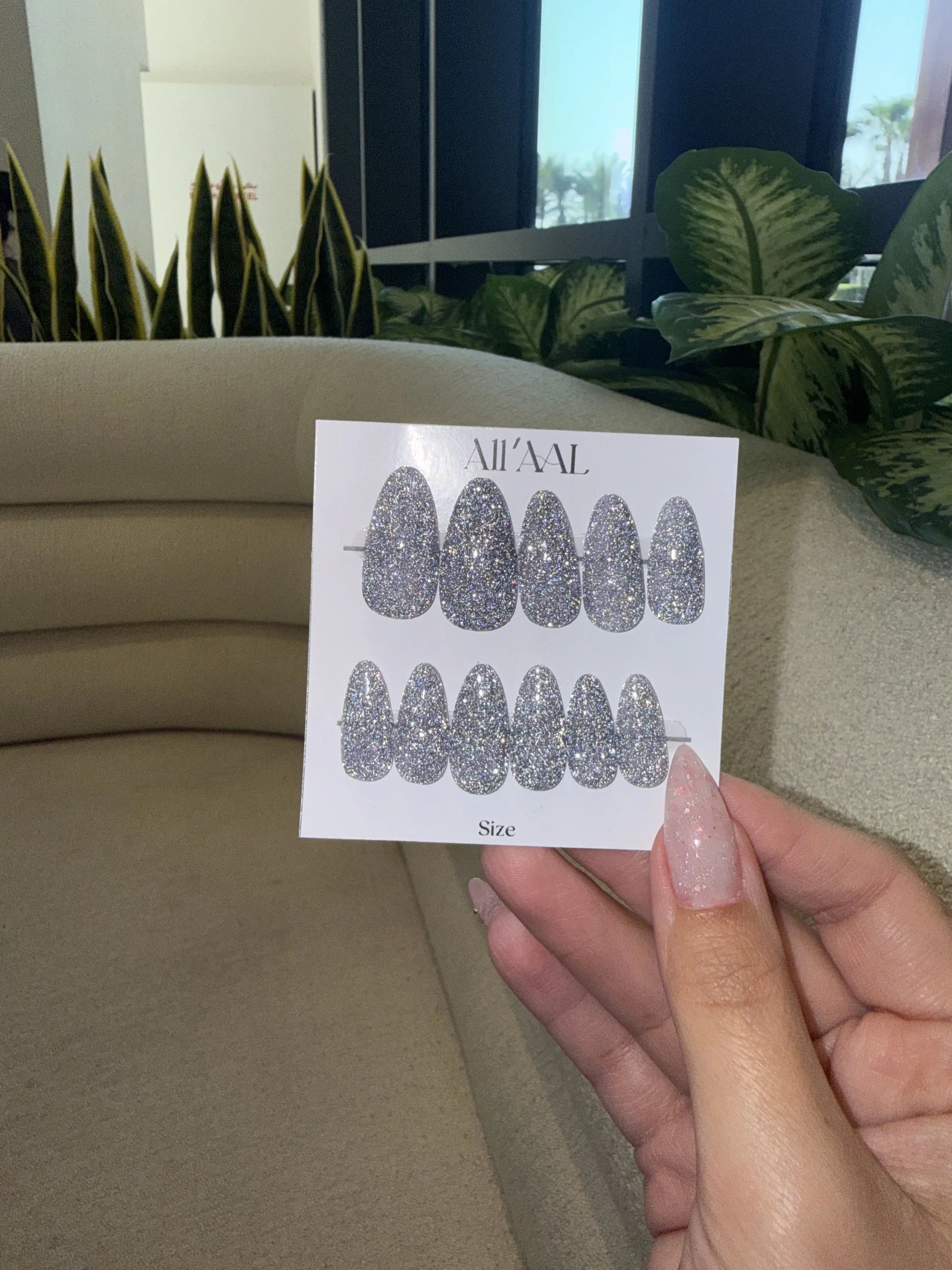 Hand holding a card with glittery nail designs in front of a beige sofa and plants.