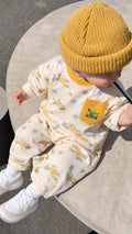 A baby wearing a mustard yellow knit beanie, a cream-colored jumpsuit with a yellow dinosaur print and a yellow chest pocket, and white sneakers, sitting on a round concrete surface.