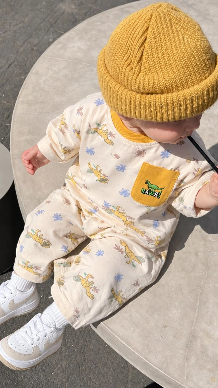A baby wearing a mustard yellow knit beanie, a cream-colored jumpsuit with a yellow dinosaur print and a yellow chest pocket, and white sneakers, sitting on a round concrete surface.