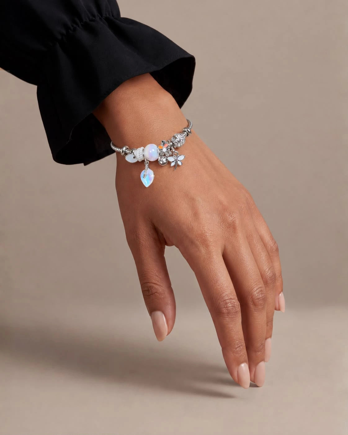 A close-up of a person's hand wearing a delicate silver charm bracelet with white and light blue stones, set against a solid beige background.